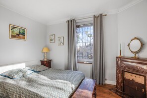 A bedroom with a bed, a window with curtains, a bedside table with a lamp, a mirror, and a fireplace.