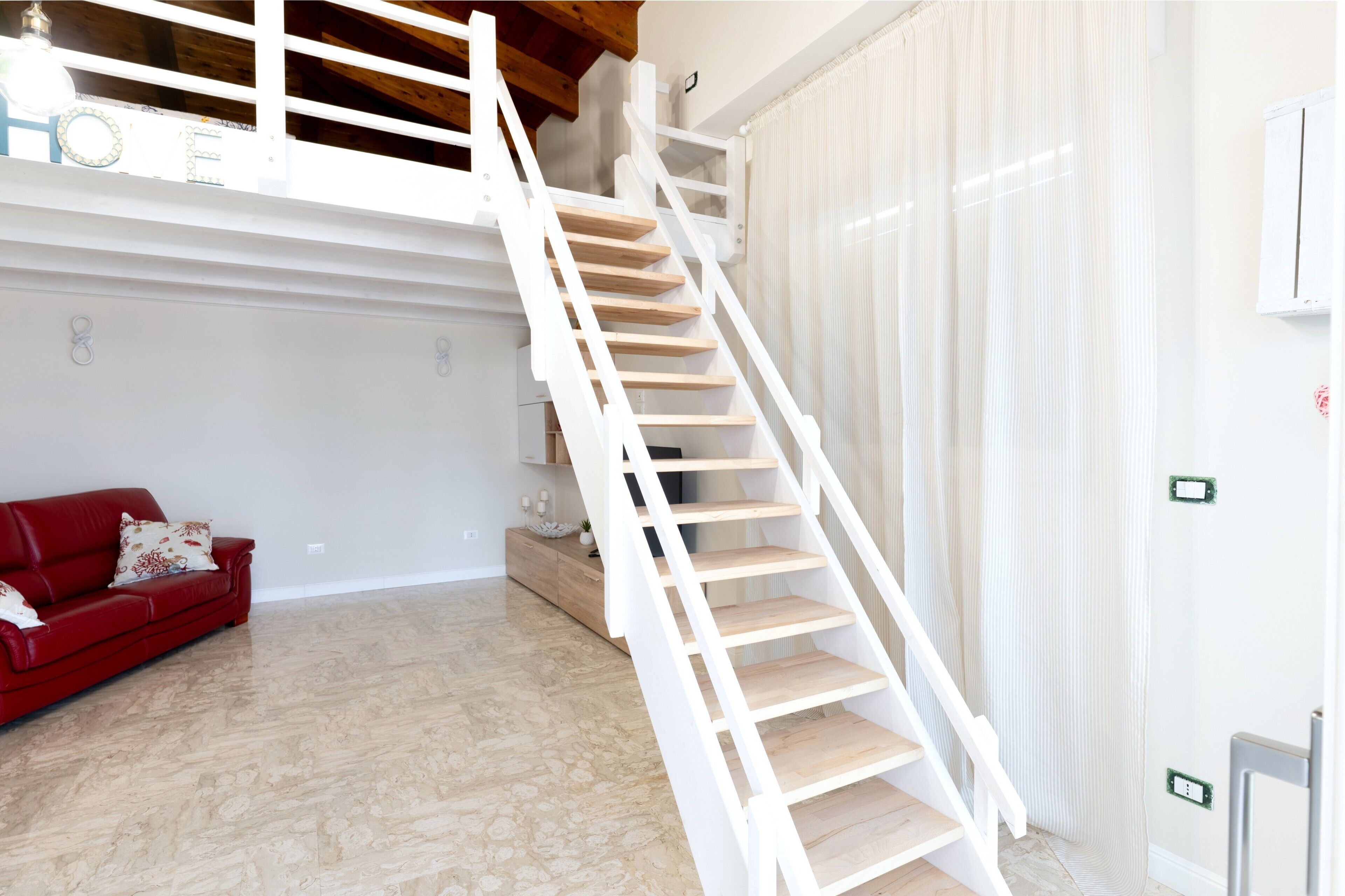 Un escalier avec des rampes blanches et des marches en bois, un canapé rouge et un tapis à motifs dans une pièce aux murs blancs et au plafond en bois.