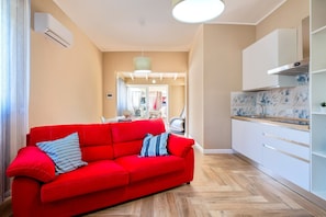 A modern living room with a red sofa, a kitchen with white cabinets, and a herringbone wood floor.