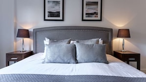 A neatly made bed with grey bedding and pillows, flanked by two bedside tables with lamps, and framed pictures on the wall. - Spacious 2-Bedroom Executive Apartment – Ideal for Business Travellers (London)