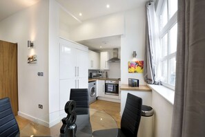 A compact kitchen with white cabinets, a microwave, oven, and a washing machine. There is a dining area with a glass table and black chairs. - Premium Serviced Apartments in Barbican, London – Perfect for Short and Corporate Clients (London)