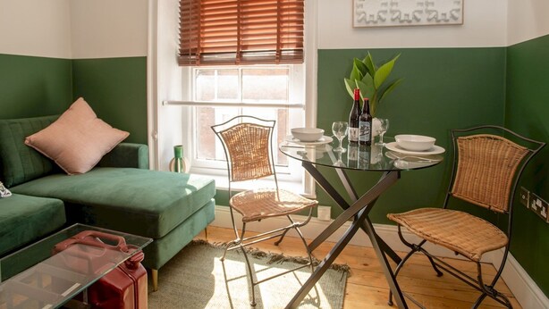 Een ruimtelijk ingerichte leefruimte met een groene bank, een glazen eettafel met stoelen en een raam met zonwering.