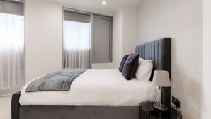 A modern bedroom with a large bed, a bedside table with a lamp, and a window with blinds. - Modern 1-Bedroom Stay in Stratford near to Canary Wharf | Ideal for Business or Leisure (London)