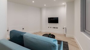 A modern living room with a teal sofa, a flat-screen TV mounted on the wall, a small round coffee table, and a window on the right. - Modern 1-Bedroom Stay in Stratford near to Canary Wharf | Ideal for Business or Leisure (London)