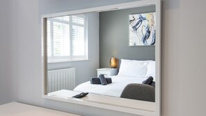 Una habitación de hotel moderna con un espejo grande, una cama con sábanas blancas, una silla gris, una mesita de noche con una lámpara y una ventana con persianas.