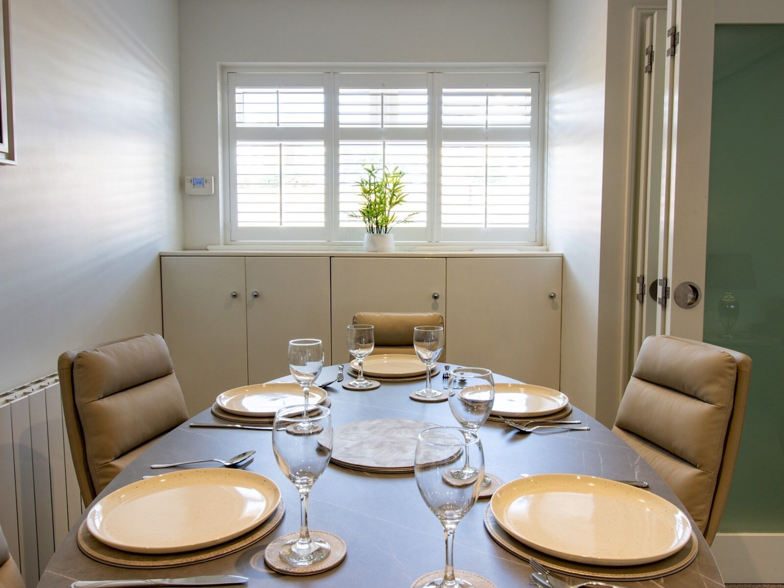 A dining area with a table set for four, beige chairs, and a window with blinds.