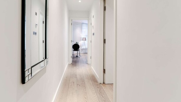 Un couloir étroit aux murs blancs et au plancher en bois, qui mène à une pièce où se trouvent une table ronde et une lampe.