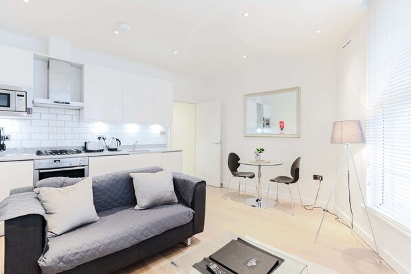 A modern kitchen with white cabinets, a microwave, and a stove. There is a grey sofa with cushions in the living area, and a small round table with two chairs. A lamp and a mirror are on the wall. - Prime Serviced Apartments in Trafalgar Square, London (London)