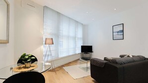 Un salon moderne avec un canapé gris foncé, une table basse en verre ornée d’une plante en pot, un lampadaire et un téléviseur à écran plat.