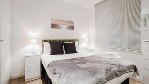 Une chambre à coucher moderne avec un grand lit, deux tables de chevet avec des lampes, une tête de lit à motifs rayés, un tableau au mur et une fenêtre avec des stores.
