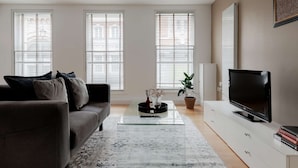 A modern living room with a grey sofa, a glass coffee table, a TV on a white cabinet, and three large windows with blinds. - Premium Serviced Apartments in Covent Garden (London)