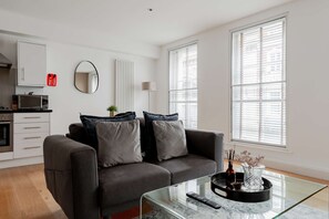 A modern living room with a grey sofa, a glass coffee table, and a kitchen area with a microwave.