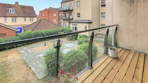A balcony with a wooden floor and glass railing overlooking a courtyard with a small garden and a brick building. - Pass the Keys | Home & Haven Canterbury (Canterbury)