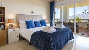 A hotel room with a large bed, a bedside table with a lamp, a bookshelf, and a view of the outdoors through a sliding glass door. - Golfer’s Paradise! Spacious Apt in La Cala Golf | Pass the Keys (Mijas)