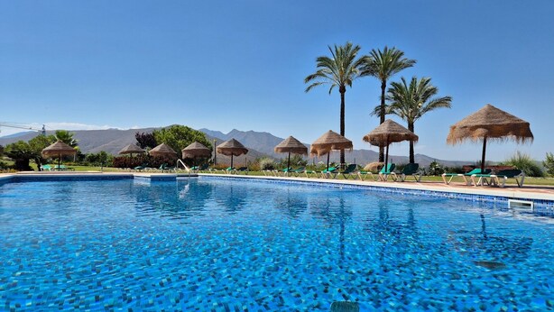A swimming pool with clear blue water, surrounded by lounge chairs and thatched umbrellas, with mountains in the background. - Golfer’s Paradise! Spacious Apt in La Cala Golf | Pass the Keys (Mijas)