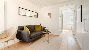 Un salon moderne avec un canapé gris, une petite table d’appoint et un fauteuil. Un miroir est accroché au mur, et une porte donne accès à une autre pièce.
