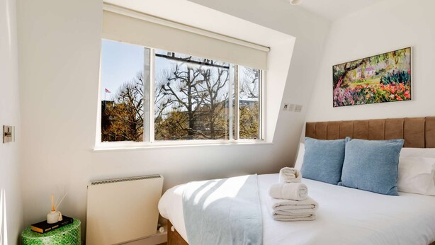 Une chambre avec une grande fenêtre, un radiateur, une table de chevet avec un livre, un lit avec une literie blanche et des oreillers bleus, et un tableau accroché au mur.