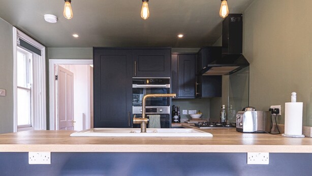 A modern kitchen with dark cabinets, a stainless steel range, and a wooden countertop. There are three wooden stools in front of the counter.