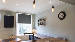 A modern kitchen with a central island, pendant lighting, a window with blinds, a clock, and a plant shelf.
