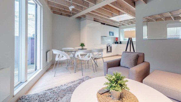 Una sala moderna con un sofá, una mesa de centro con una planta, y un comedor con sillas blancas y una mesa de vidrio.