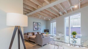 Una sala de estar moderna con un sofá, una mesa de vidrio y un televisor. Cuenta con un ventanal grande y un techo con vigas a la vista.