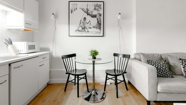 A modern kitchen with white cabinets, a microwave, and a dining area with two chairs and a small table. A framed black and white photo hangs on the wall above the dining area. - Luxurious Serviced Apartments in the Heart of Marylebone, London (London)