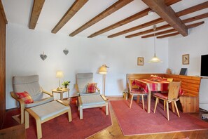 Ein gemütlicher Raum mit Holzbalken, einem Sofa, Sesseln, einem Esstisch und einem Fernseher.