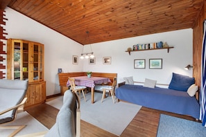 A cozy room with a wooden ceiling, a bed with a blue polka dot bedspread, a wooden cabinet, a dining table with a checkered tablecloth, and a chair.
