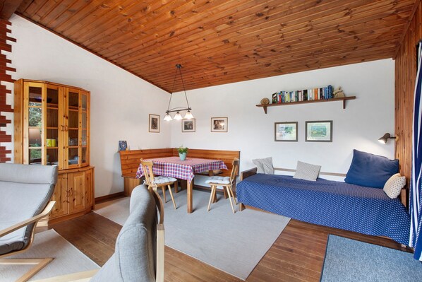 A cozy room with a wooden ceiling, a bed with a blue polka dot bedspread, a wooden cabinet, a dining table with a checkered tablecloth, and a chair.