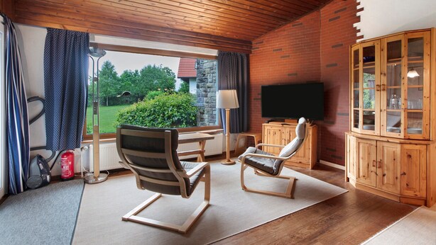 Une pièce avec un plafond en bois, une télévision, un meuble en bois, une chaise, une table et une vue sur la verdure à l’extérieur.