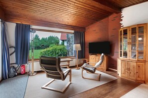 Una habitación con techo de madera, televisor, aparador de madera, una silla, una mesa y vista a espacios verdes.