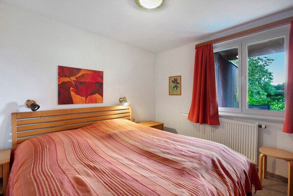 Un dormitorio con cama, mesita de noche, ventana con cortinas y radiador.