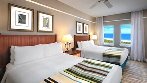 A hotel room with two beds, a wooden headboard, a ceiling fan, framed artwork on the wall, and a view of the ocean.