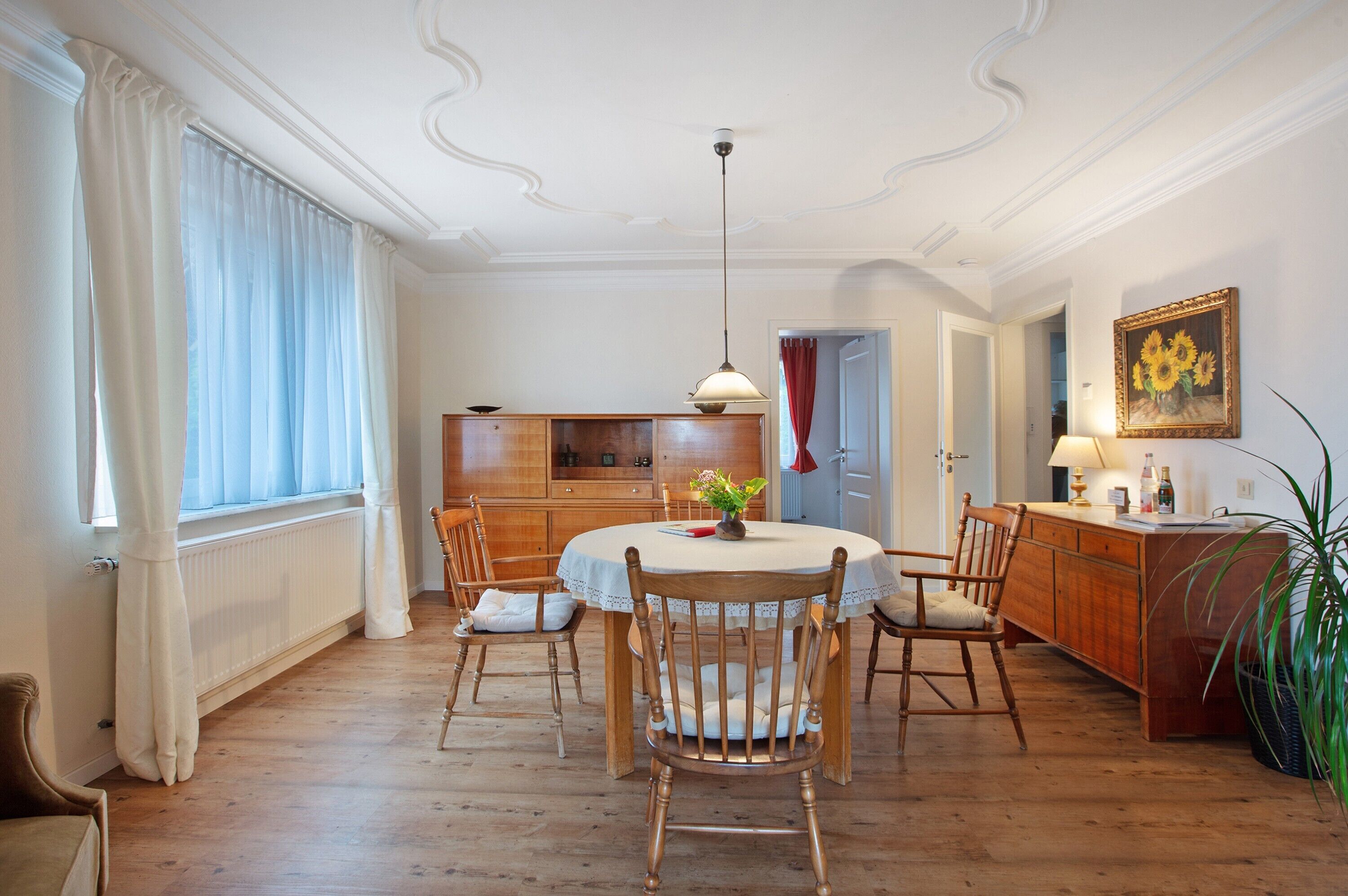 Et spiseområde med et rundt bord, stole, en hængende lampe og et køkken i baggrunden.