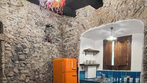 A rustic kitchen with a stone wall, a wooden ceiling, a vintage orange refrigerator, a brown sofa, a tiled floor, and a glass table with a chair.