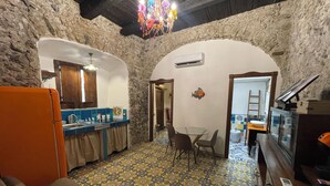 A rustic kitchen with a stone wall, a wooden ceiling, and a colorful chandelier. There is a dining table with chairs and a small TV on a wooden table.