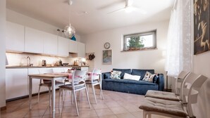 Une cuisine moderne avec des meubles blancs, une table à manger avec des chaises, un canapé bleu avec des coussins à motifs et une fenêtre donnant sur des arbres.