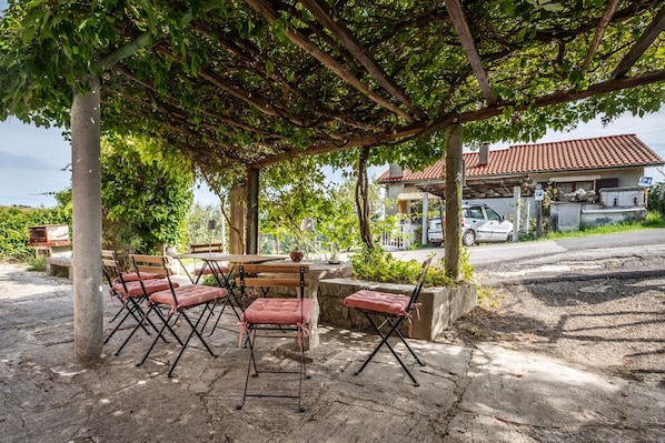 Ein Außenbereich mit Tischen und Stühlen, einer mit Wein bewachsenen Pergola und einem Gebäude im Hintergrund.