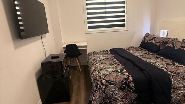 A bedroom with a bed, a TV, a window with blinds, a desk, and a fluffy rug. - Serenity Space (Niagara Falls)