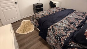 A bedroom with a bed, a TV, a desk, and a window with blinds. - Serenity Space (Niagara Falls)