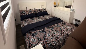 A bedroom with a bed, a dresser, a mirror, and a window with blinds. - Serenity Space (Niagara Falls)