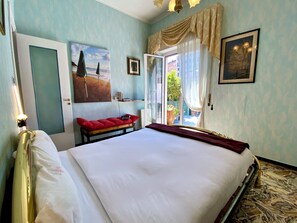 Una habitación de hotel con una cama, un banco con cojines rojos, un cuadro, una ventana con cortinas y una puerta.