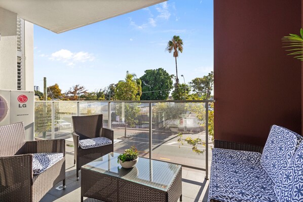 A balcony with wicker furniture, a glass table with a plant, and a view of the street and trees. - Beaufort Street 1bdr Parking (Perth)
