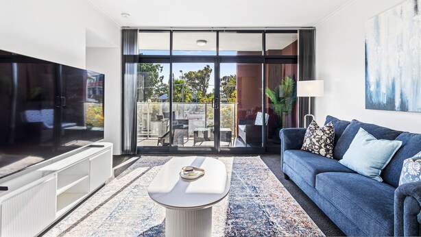 A modern living room with a flat-screen TV, a sofa with cushions, a coffee table, and a large sliding glass door leading to an outdoor patio area. - Beaufort Street 1bdr Parking (Perth)
