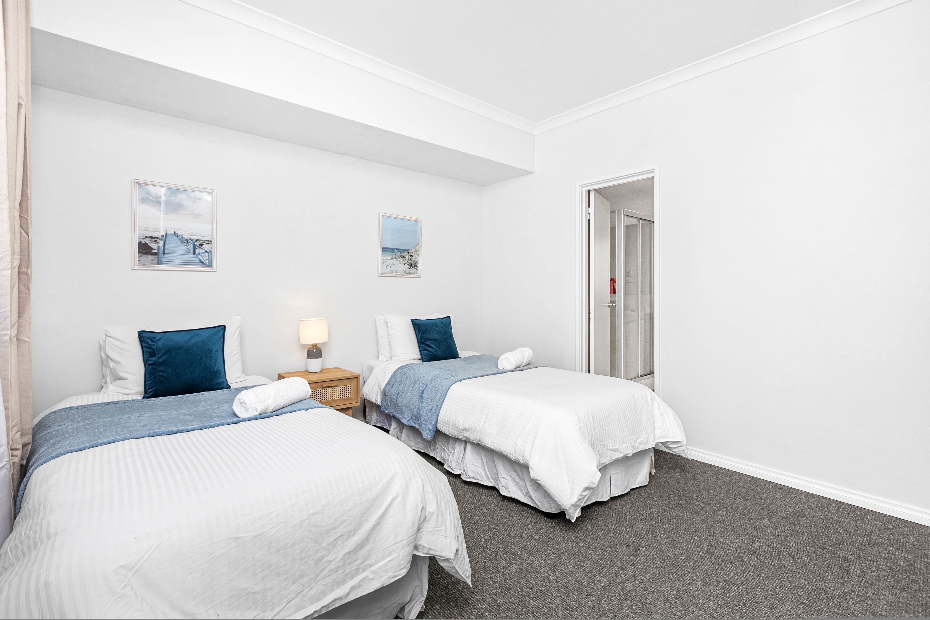 A hotel room with two single beds, a nightstand with a lamp, and two framed pictures on the wall.