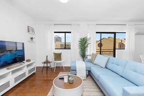 Ein modernes Wohnzimmer mit einem großen Flachbildfernseher, einem hellblauen Sofa, einem Couchtisch und Blick auf Gebäude durch große Fenster.