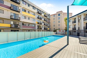 A swimming pool with a wooden deck, surrounded by multi-story residential buildings with balconies. - Modern 2BR Pool Gym Parking (Perth)