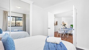 A hotel room with two beds, a dining area with a table and chairs, and a view of buildings outside. - Modern 2BR Pool Gym Parking (Perth)