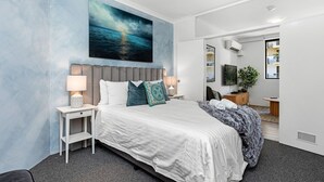 A modern hotel room with a large bed, a nightstand with a lamp, a painting of a seascape, and a view of the city through an open door. - Stunning 1bdr Paradise - Gym Pool (Perth)