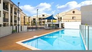 A swimming pool with a wooden deck, surrounded by buildings with balconies and a covered seating area. - Stunning 1bdr Paradise - Gym Pool (Perth)